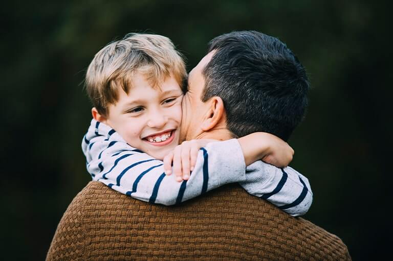 Young boy with blonde hair and a stripey jumper hugging man with black hair and brown jumper